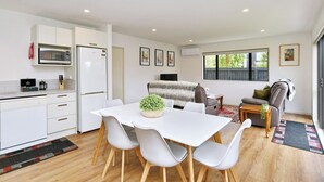 A modern kitchen with white cabinetry, a microwave, and a dining area with a white table and chairs. There is a small plant on the table, and the room has a large window with a view of trees. - Brand-new, townhouse - walking distance to Westfield Mall (Christchurch)