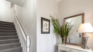 Una escalera con barandillas blancas, una obra de arte enmarcada en la pared, un florero con follaje y una lámpara sobre una consola.