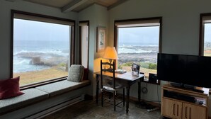 A room with a large window overlooking the ocean, a wooden table with a lamp, a chair, and a television on a wooden stand. - NEW LISTING! Fantasea! Enjoy dramatic crashing waves, fireplace, dog friendly! (Yachats)