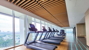 Un gimnasio con cintas de correr alineadas a lo largo de una pared con amplios ventanales que ofrecen vistas panorámicas de la ciudad.