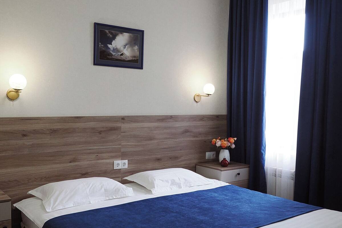 Ein Hotelzimmer mit einem hölzernen Kopfteil, einem großen Bett mit weißer und blauer Bettwäsche, zwei Nachttischen mit Lampen, einem gerahmten Bild an der Wand und einem Fenster mit dunklen Vorhängen.