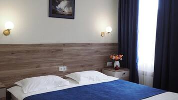 Une chambre d’hôtel avec une tête de lit en bois, un grand lit avec une literie blanche et bleue, deux tables de chevet avec des lampes, un tableau encadré au mur et une fenêtre avec des rideaux foncés.