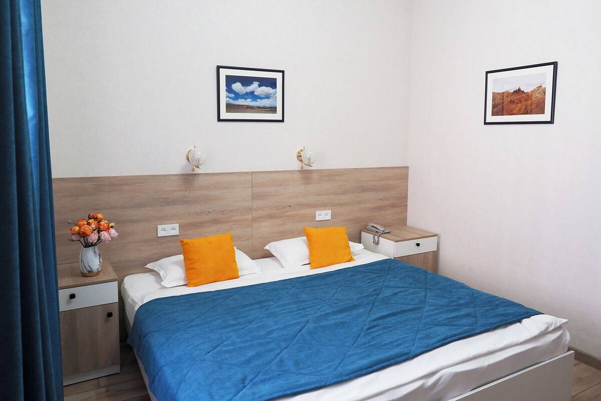 Ein ordentlich eingerichtetes Hotelzimmer mit einem Bett, zwei Nachttischen, einer Lampe und einer Blumenvase sowie zwei gerahmten Bildern an der Wand.