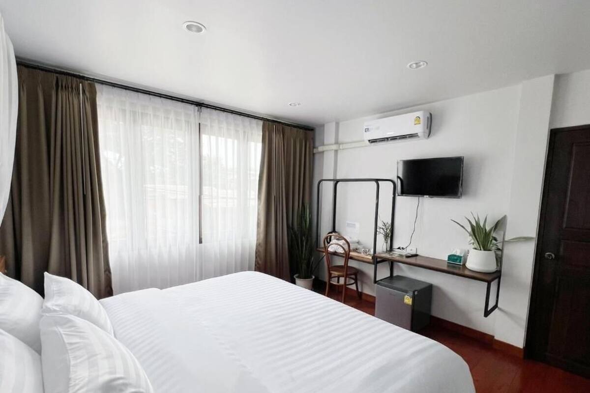 A hotel room with a bed, a desk, a chair, a TV, and an air conditioning unit.