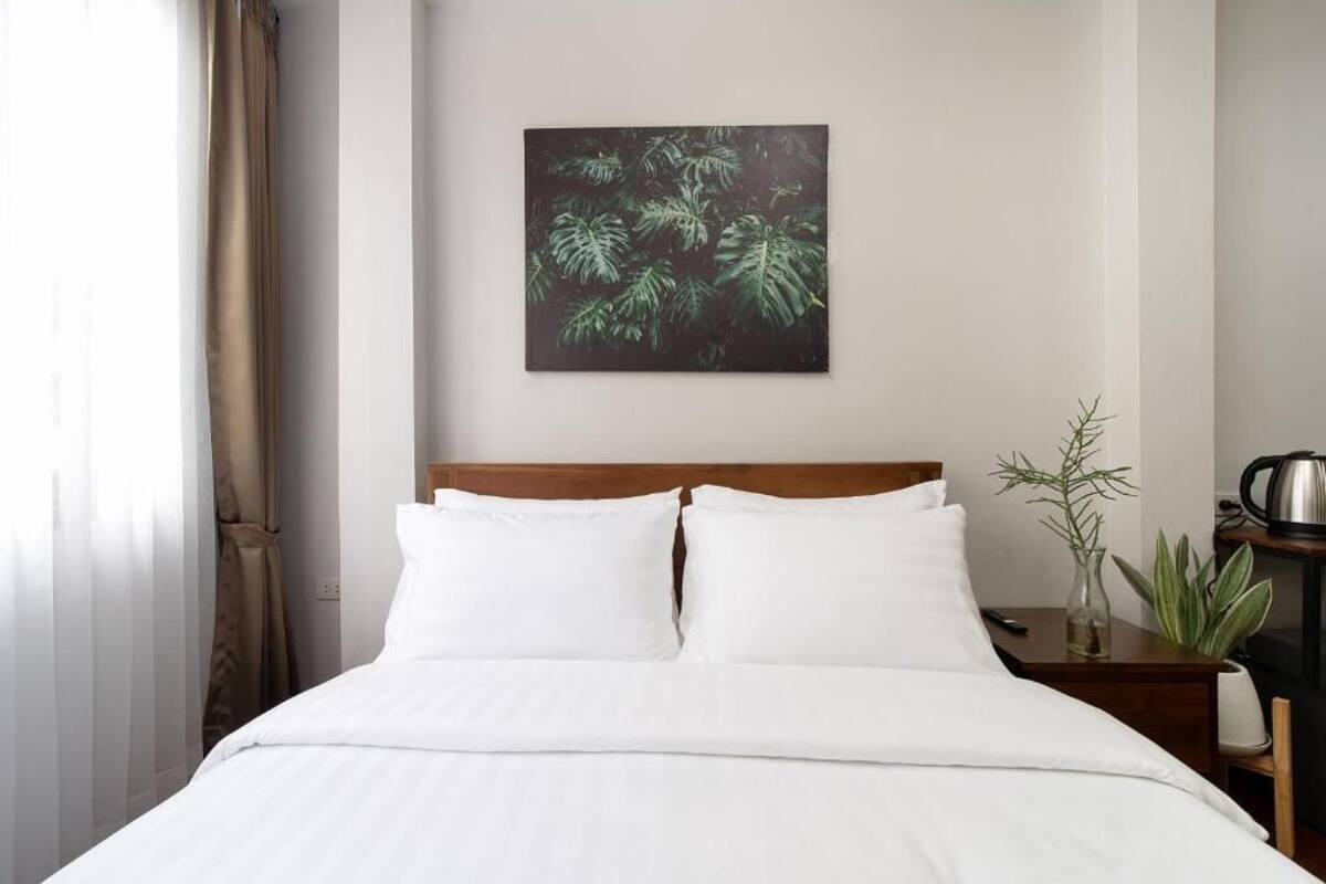 A neatly made bed with white linens, a wooden headboard, and a wall-mounted artwork of green leaves.