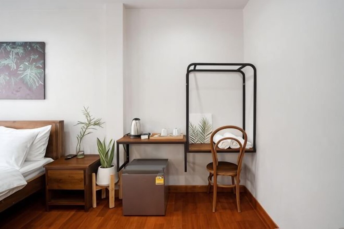 A small, neatly arranged room with a bed, bedside table, a desk with a chair, a small refrigerator, and a wall-mounted rack.
