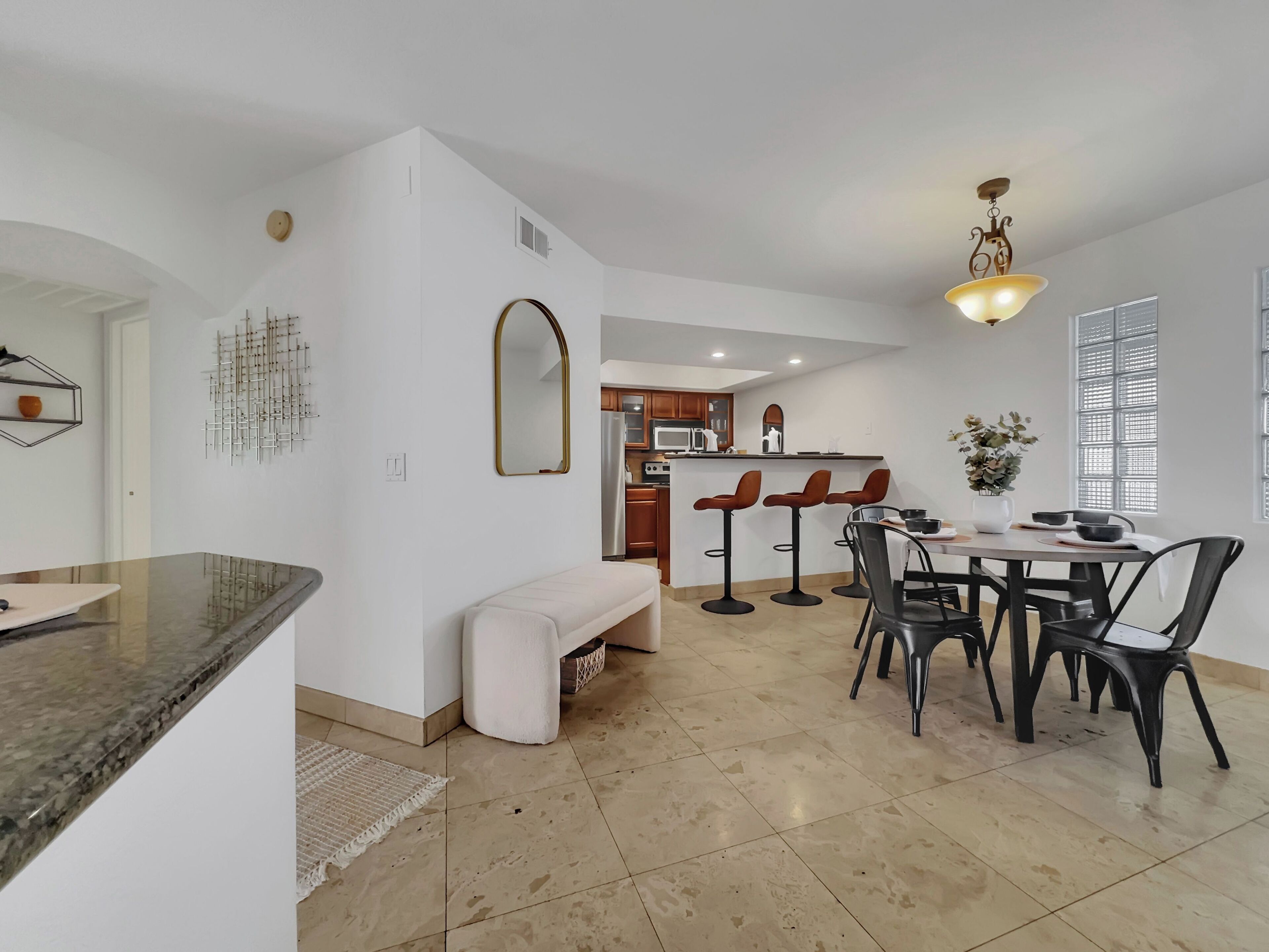 A modern kitchen with a central island, a dining area with bar stools, and a large window with blinds.