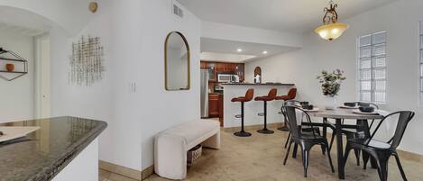 A modern kitchen with a central island, a dining area with bar stools, and a large window with blinds.