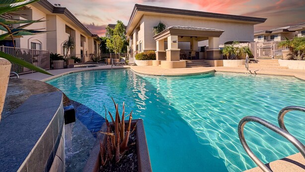 A swimming pool with a covered seating area, surrounded by buildings with a patio and a gate.