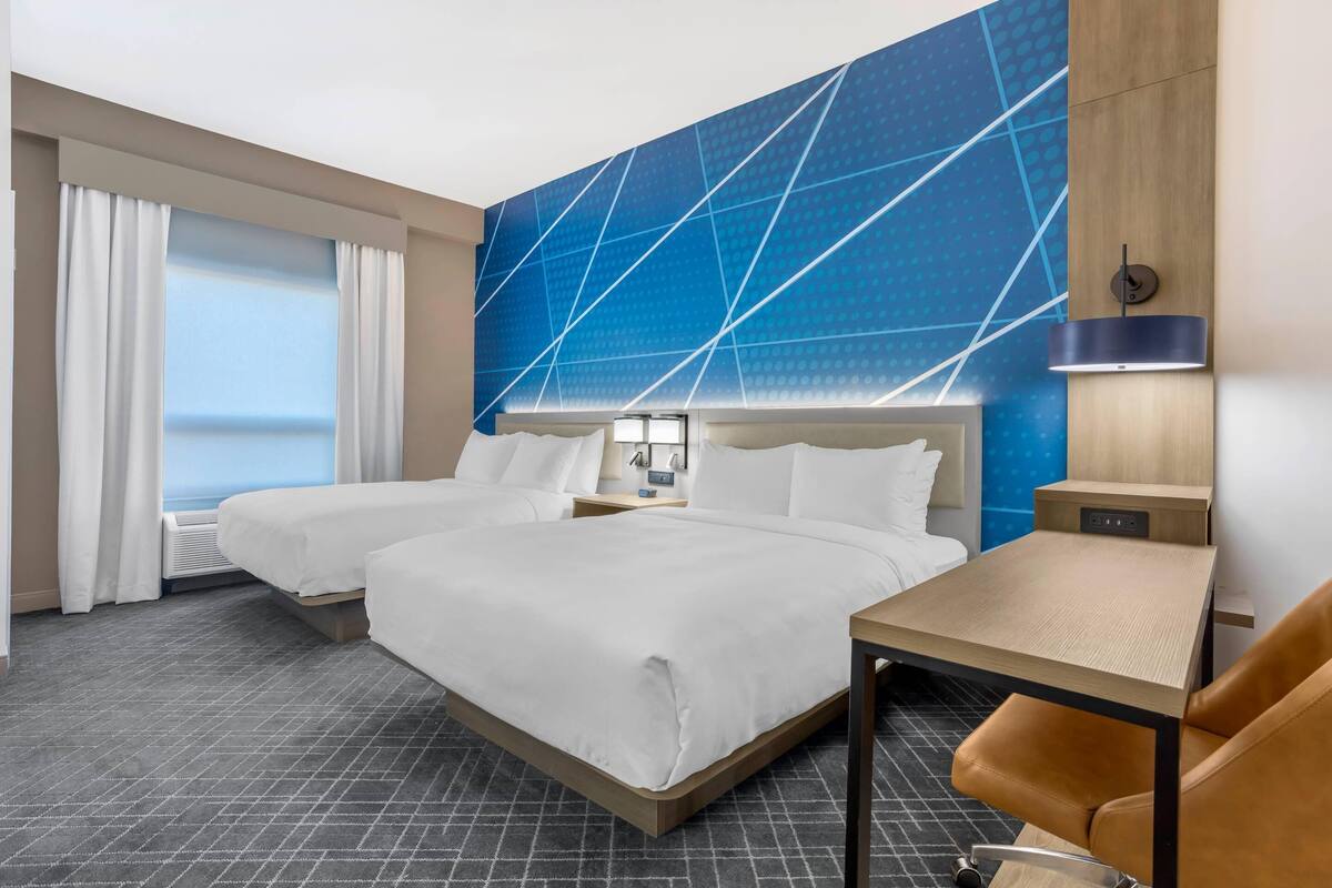 Hotelkamer met twee bedden, een bureau, een stoel, een groot raam met gordijnen en een blauw geometrisch wandontwerp.
