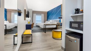 A modern hotel room with a kitchenette, a bed with white bedding, a seating area with a yellow ottoman, and a large window with curtains. - Comfort Suites Central Valley - Woodbury (Central Valley)