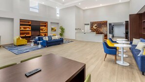 A spacious living area with a blue sofa, yellow armchair, and a coffee table. There is a TV mounted on a wooden shelf, and a kitchen area is visible in the background. - Comfort Suites Central Valley - Woodbury (Central Valley)