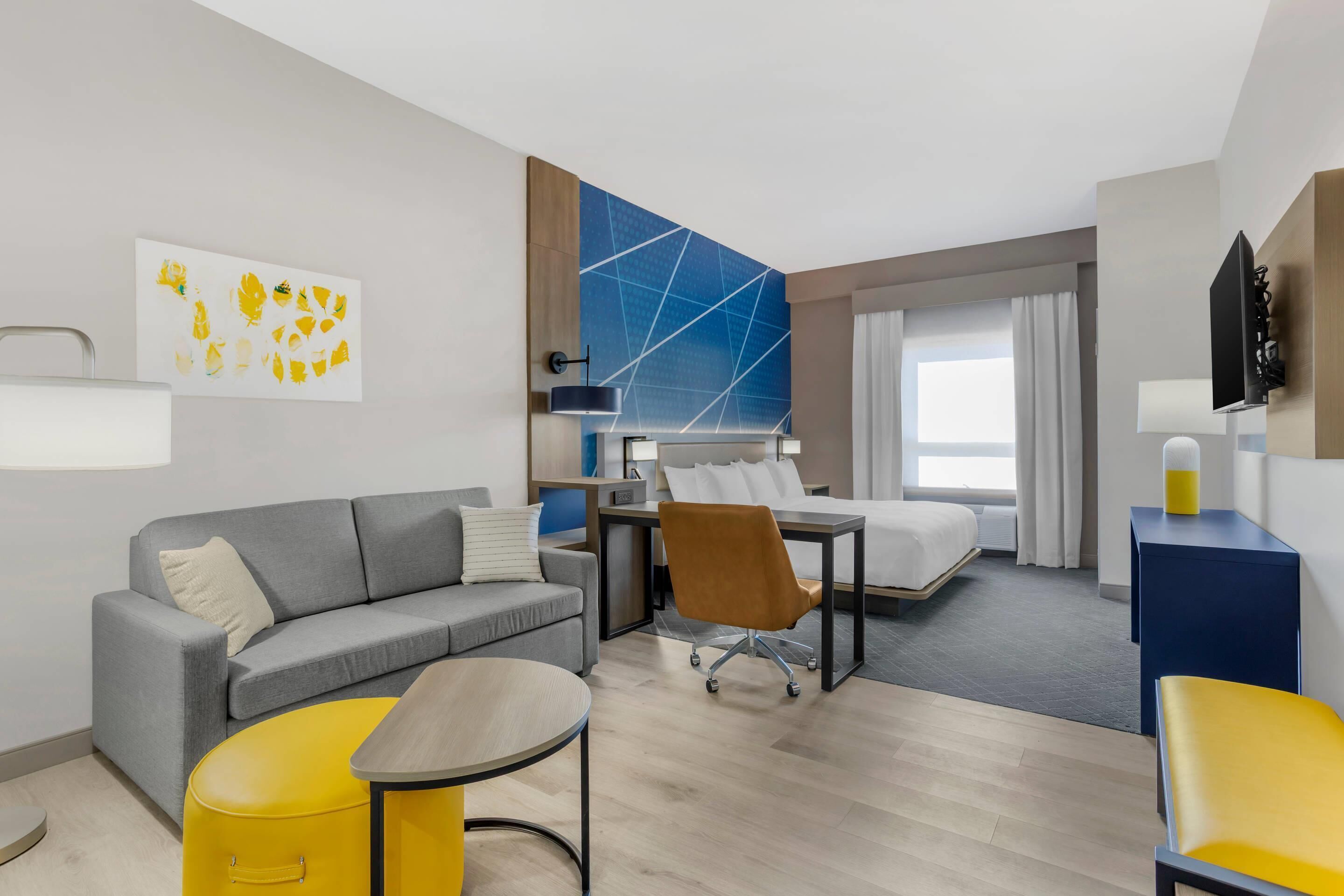 Una habitación de hotel moderna con un sofá gris, un reposapiés amarillo, un escritorio con una silla, una cama y un ventanal con cortinas.