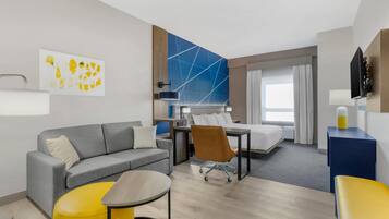 Una habitación de hotel moderna con un sofá gris, un reposapiés amarillo, un escritorio con una silla, una cama y un ventanal con cortinas.