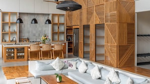 A modern interior with a wooden ceiling, a central seating area with a sofa and chairs, a kitchen with wooden cabinets, and pendant lighting. - Villa Lava (Canggu)
