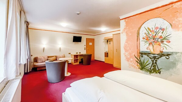 A hotel room with a bed, a sofa, a small table, a trash bin, and a painting on the wall.