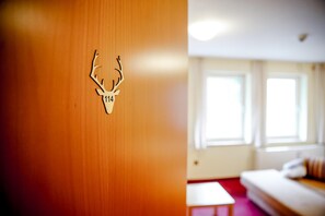 A room with a wooden wall featuring a deer head cutout and the number 114. There is a bed with a red accent wall and a window with curtains.