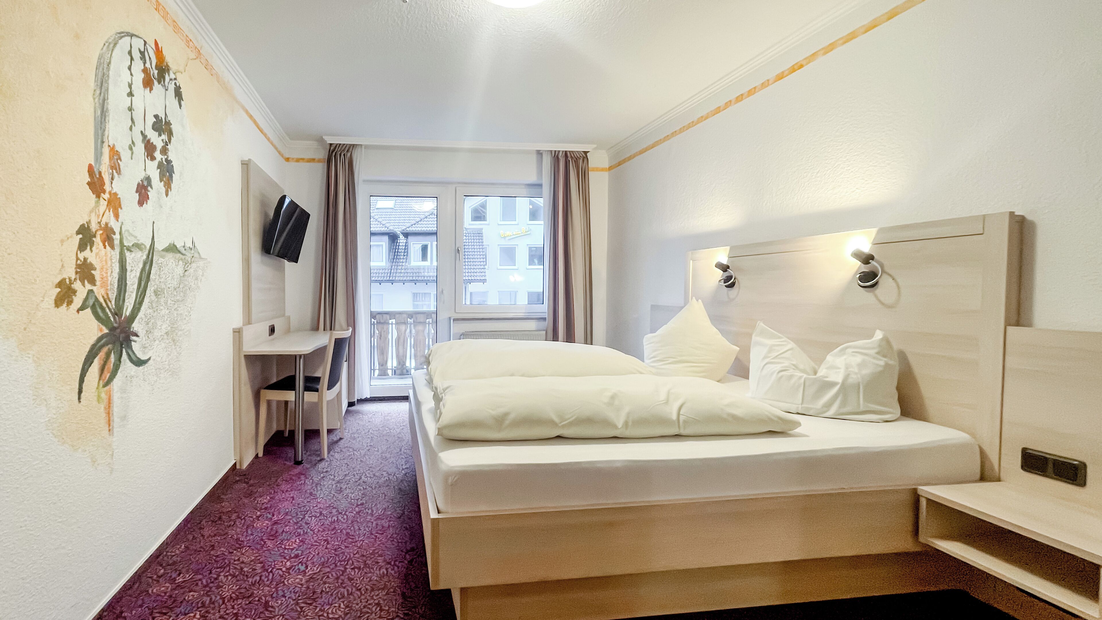 A hotel room with a bed, a desk, a TV, and a view of the outside.