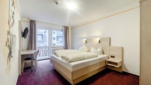 A hotel room with a bed, a desk, and a chair. There is a window with curtains and a view of buildings outside.