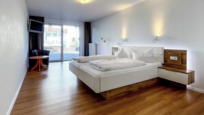 Une chambre d’hôtel moderne avec un grand lit, une table de chevet en bois, une petite table avec une chaise et une vue sur l’extérieur grâce à une porte coulissante.