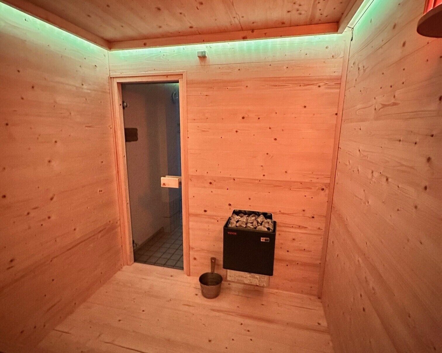 A wooden sauna room with a door, a bucket, and a box of wood pellets.