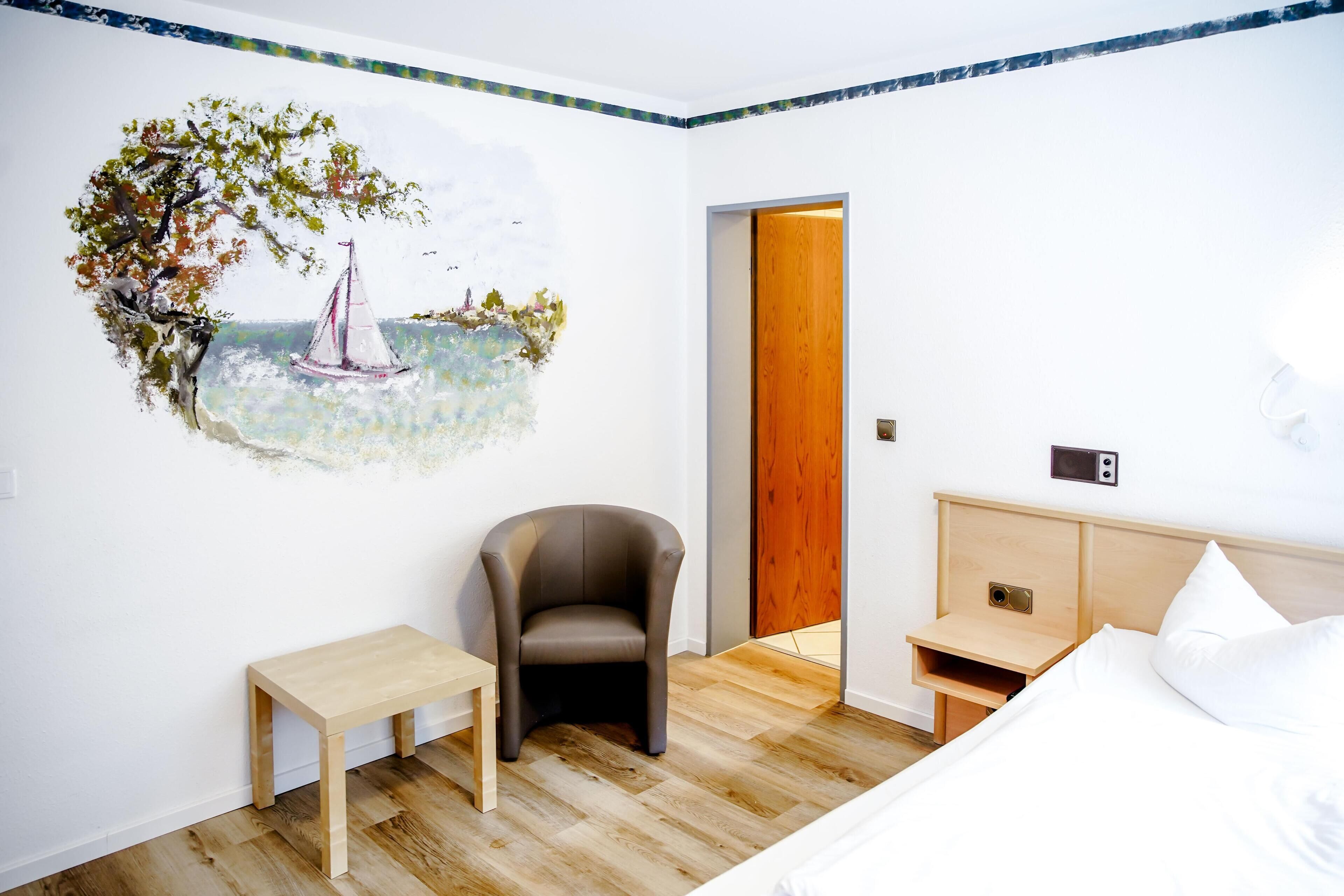 Une chambre avec un plancher en bois, un lit recouvert de draps blancs, une chaise, une petite table et une fresque murale représentant un voilier.