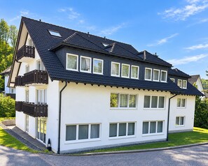 A two-story residential building with a dark roof, white exterior walls, and multiple windows. - A comfortable and versatile room—perfect for relaxation, work, and socializing (Winterberg)