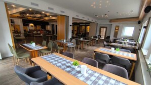 A modern dining area with long tables, chairs, and a kitchen in the background.