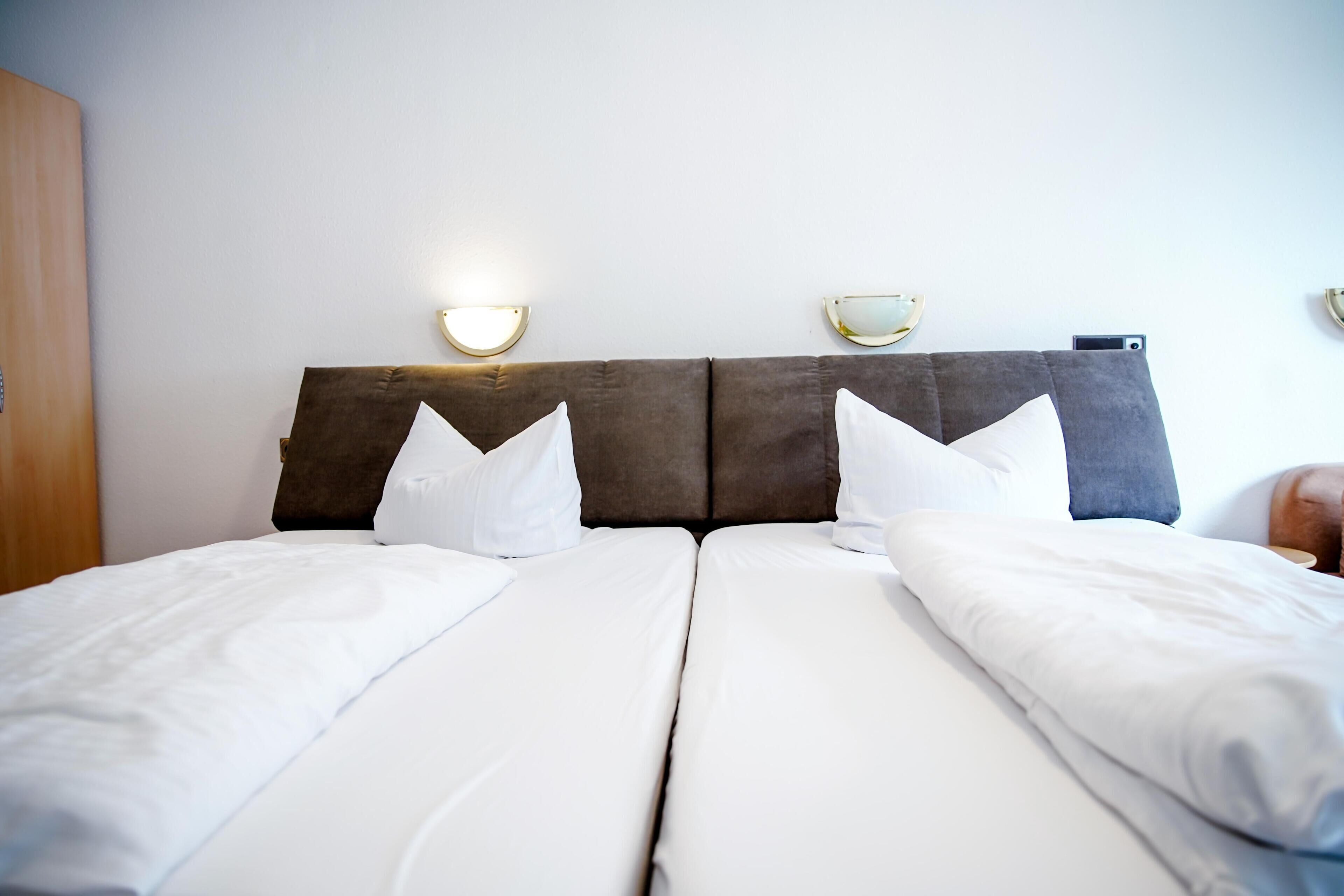 A double bed with white linens and two pillows, a wooden headboard, and wall-mounted sconces.