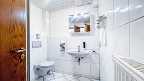 Et moderne badeværelse med hvide fliser, toilet, håndvask, spejl og bruser.
