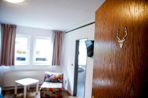 A room with a wooden door featuring a deer head emblem, a small table, a colorful chair, and a window with curtains.