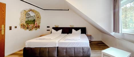 A bedroom with a large bed, a small bedside table, a bench, a mirror, and a window with curtains.