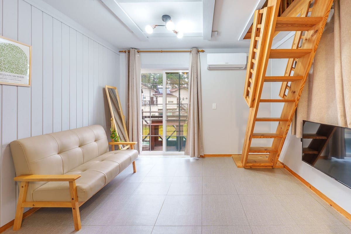 Una sala de estar con sofá, televisor, escalera de madera y un ventanal grande.
