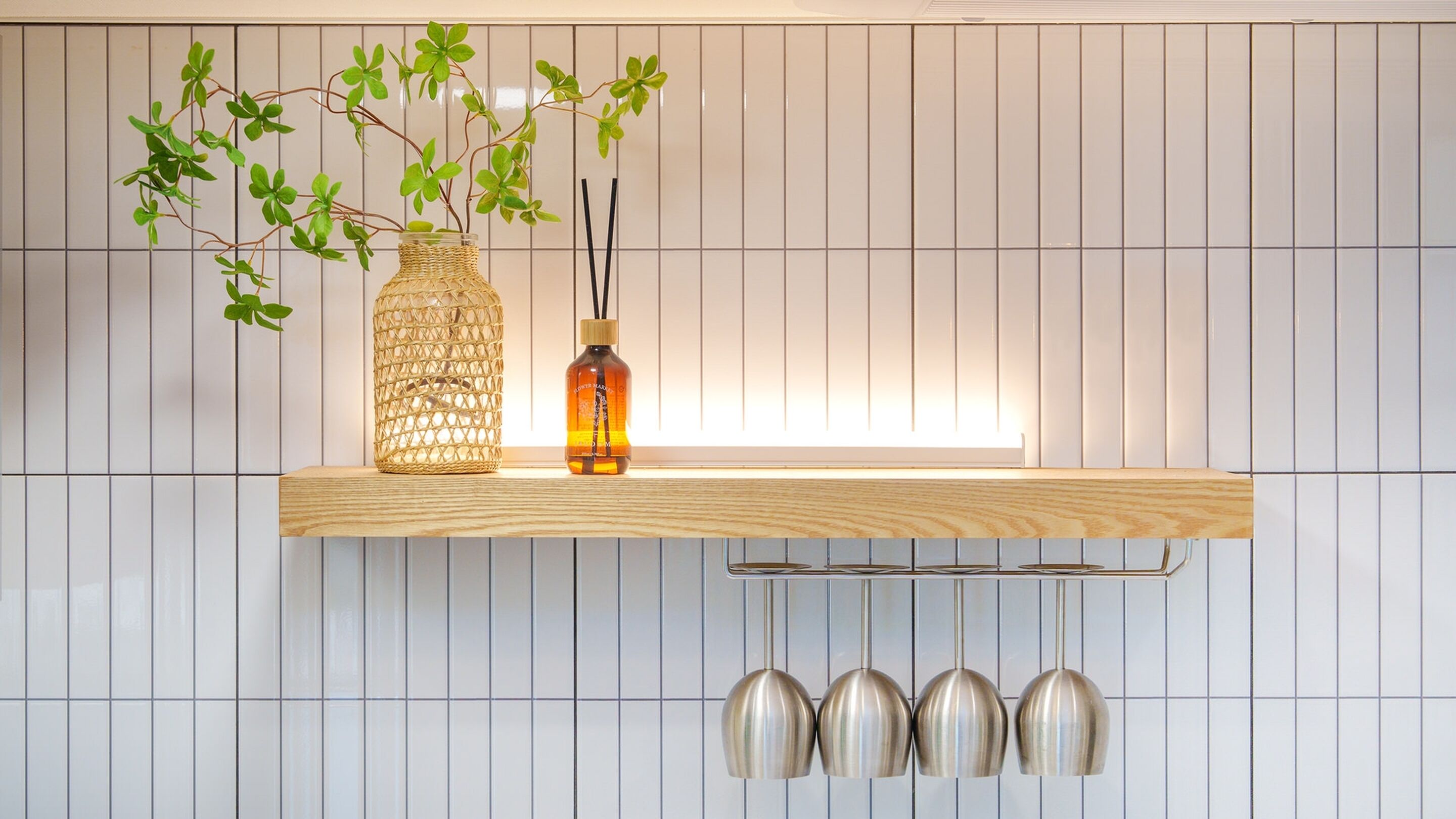 A kitchen with a wooden shelf holding a woven vase and a bottle, and a row of hanging metallic cups below.