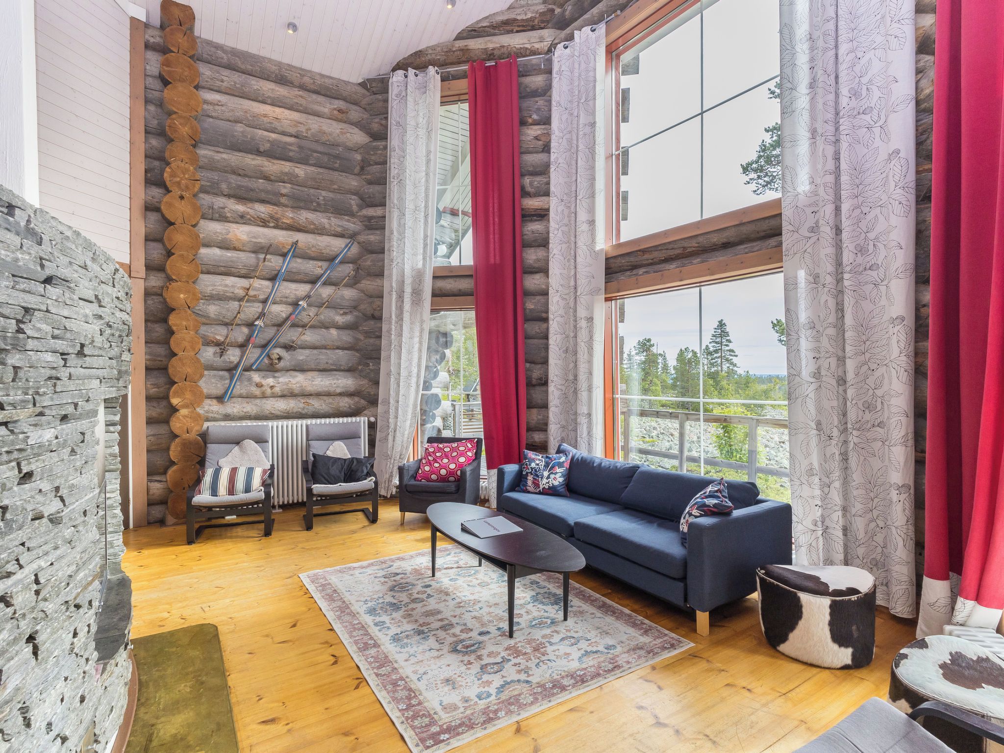 L'intérieur d'une cabane en rondins, avec un canapé, des fauteuils, un tapis et une vue sur les arbres à travers de grandes fenêtres.