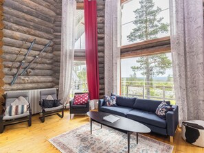 L’intérieur d’une cabane en rondins, avec un canapé, des fauteuils, une table basse et une vue sur l’extérieur.