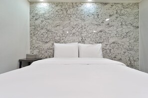 Een slaapkamer met een bed, witte beddengoed, een hoofdeinde met marmerprint en een nachtkastje met een lamp.