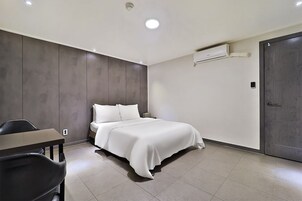 Quarto de hotel moderno com uma cama, uma escrivaninha e uma cadeira. Há um ar condicionado na parede e uma porta à direita.