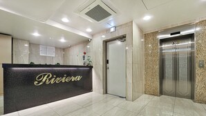 A hotel reception area with a black counter displaying the name 'Riviera'. There is a door and an elevator to the right. - Anyang Livera (Anyang)