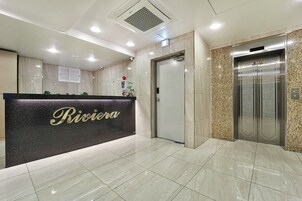 Recepção de hotel com balcão preto exibindo o nome 'Riviera'. Há uma porta e um elevador à direita.