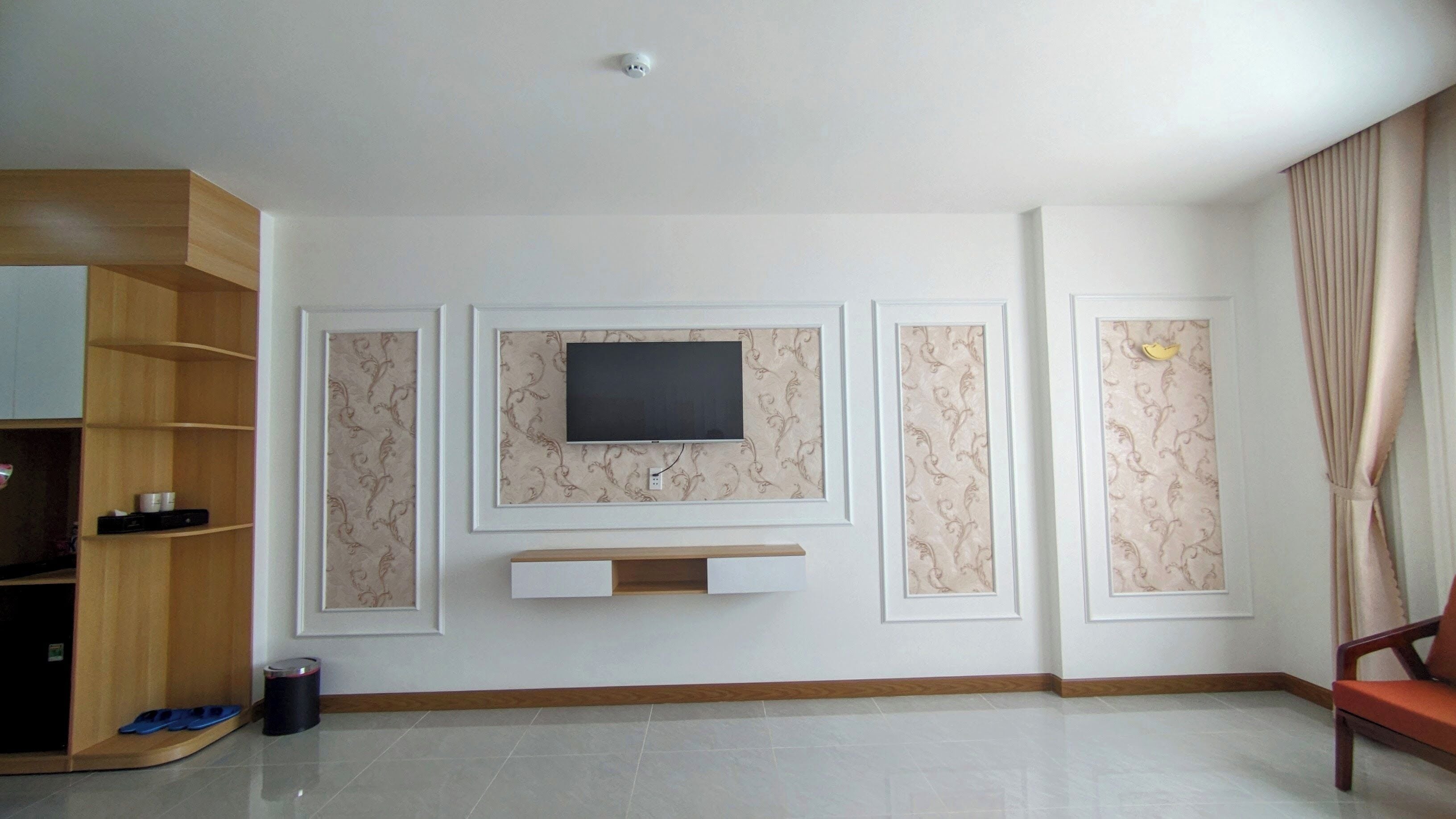 A living room with a flat-screen TV mounted on a wall with decorative paneling, a wooden entertainment unit, and a beige curtain.