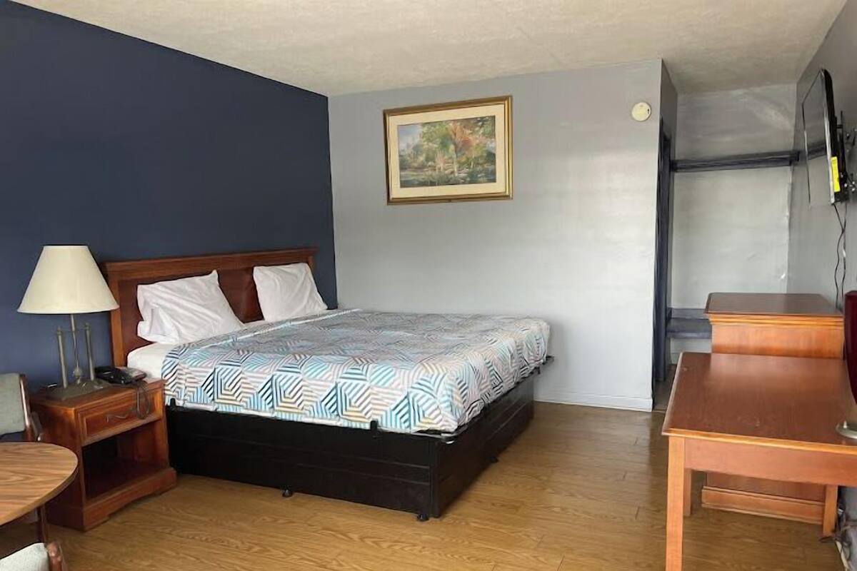 A bedroom with a bed, a nightstand, a lamp, a wooden table, and a painting on the wall.