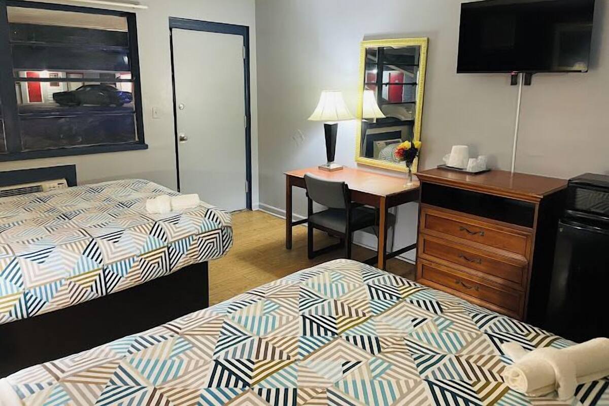 A hotel room with two beds, a desk, a chair, a dresser, a television, and a window with a view of a car.