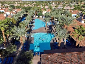 Ein Luftbild eines Resorts mit einem zentral gelegenen Swimmingpool, umgeben von Palmen und Liegestühlen.