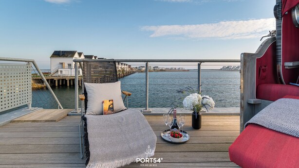 A deck with a bed, a dining table with a bottle and glasses, and a view of the water and buildings in the background.