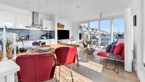 A modern kitchen with a large island, dining area with red chairs, and a living area with a red sofa and large windows overlooking a harbor.