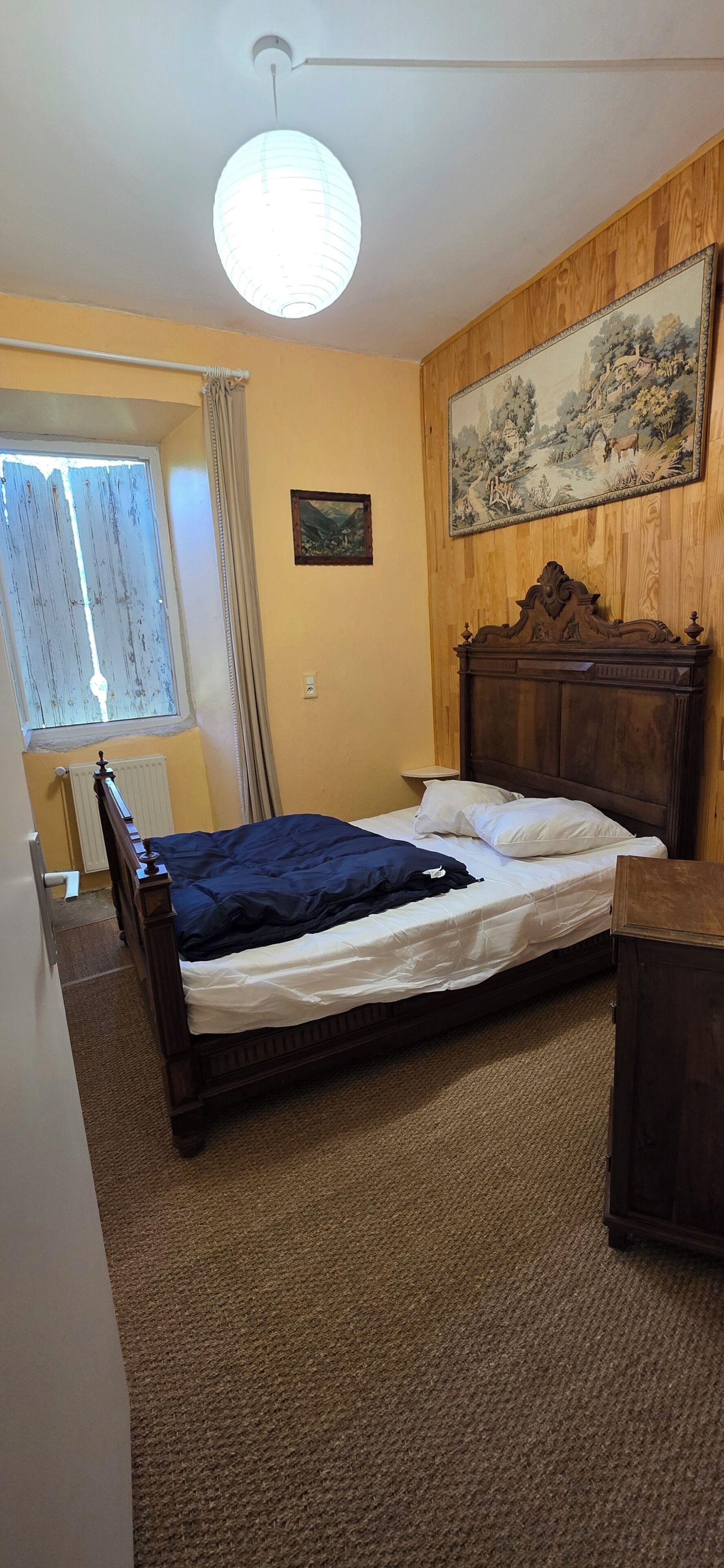 Um quarto com cabeceira de madeira, cama com roupa de cama branca e azul, uma janela com cortinas, um radiador e um quadro na parede.