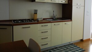 A compact kitchen with a gas stove, sink, and a dining area with a chair and table. - Olive Tree House- Vacation Rentals (Riva del Garda)
