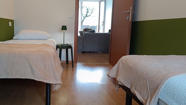 A room with two beds, a small table with a lamp, and a view of an outdoor area with a sofa. - Olive Tree House- Vacation Rentals (Riva del Garda)