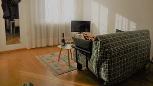 A room with a bed, a TV, a small table with a bottle and glasses, and a window with curtains. - Olive Tree House- Vacation Rentals (Riva del Garda)